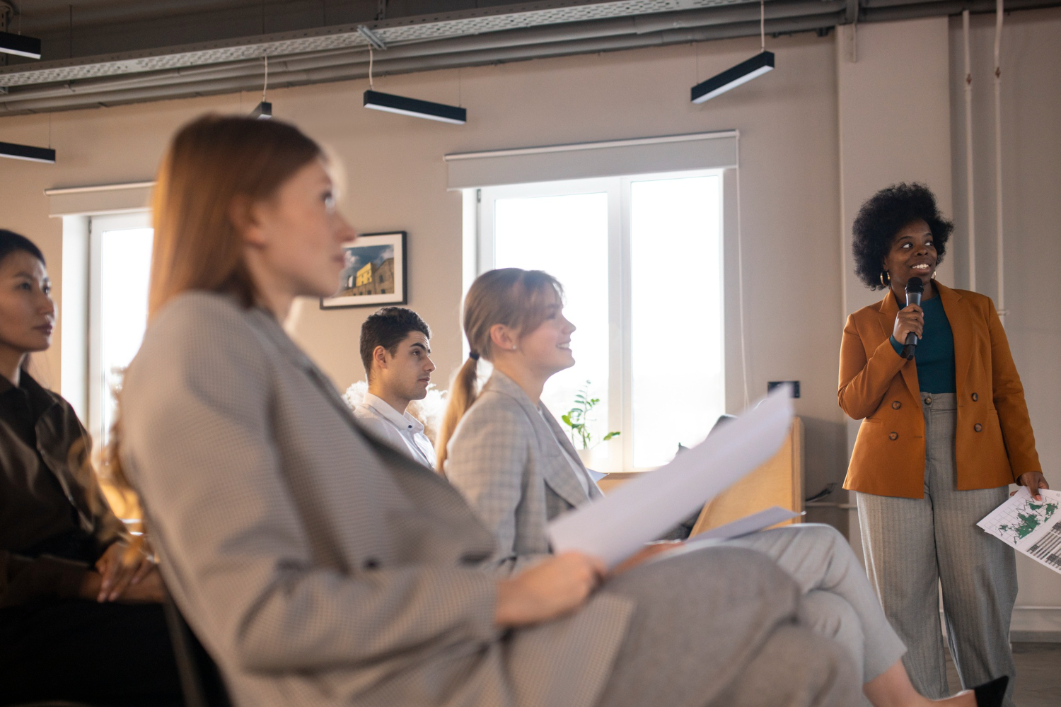 A presenter talking to 6 people in a meeting room going through merger details and the impact on related markets